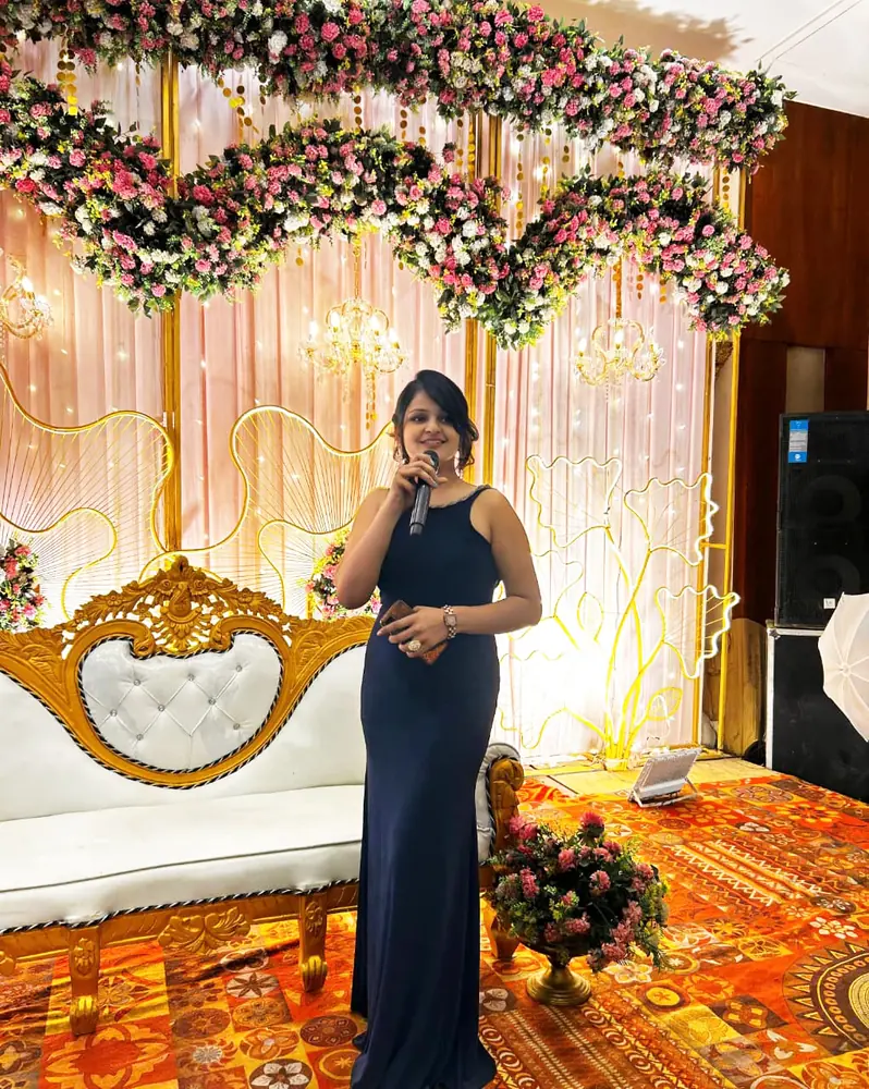 Anupriya Tripathi hosting a wedding, microphone in hand, on a floral stage